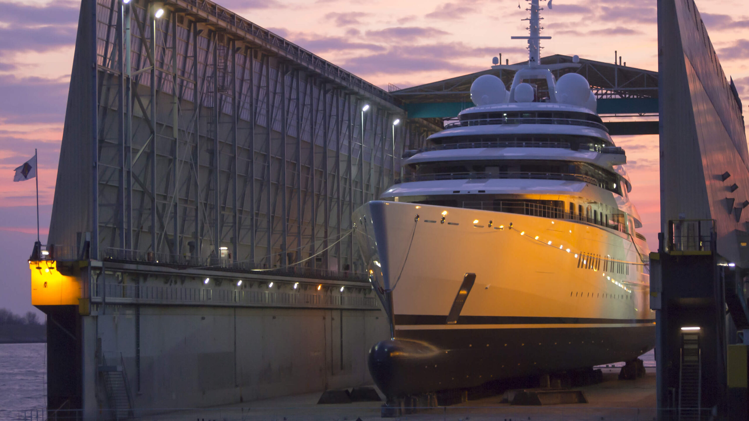 Large superyacht positioned at the entrance of a Lürssen shipbuilding hall at dusk