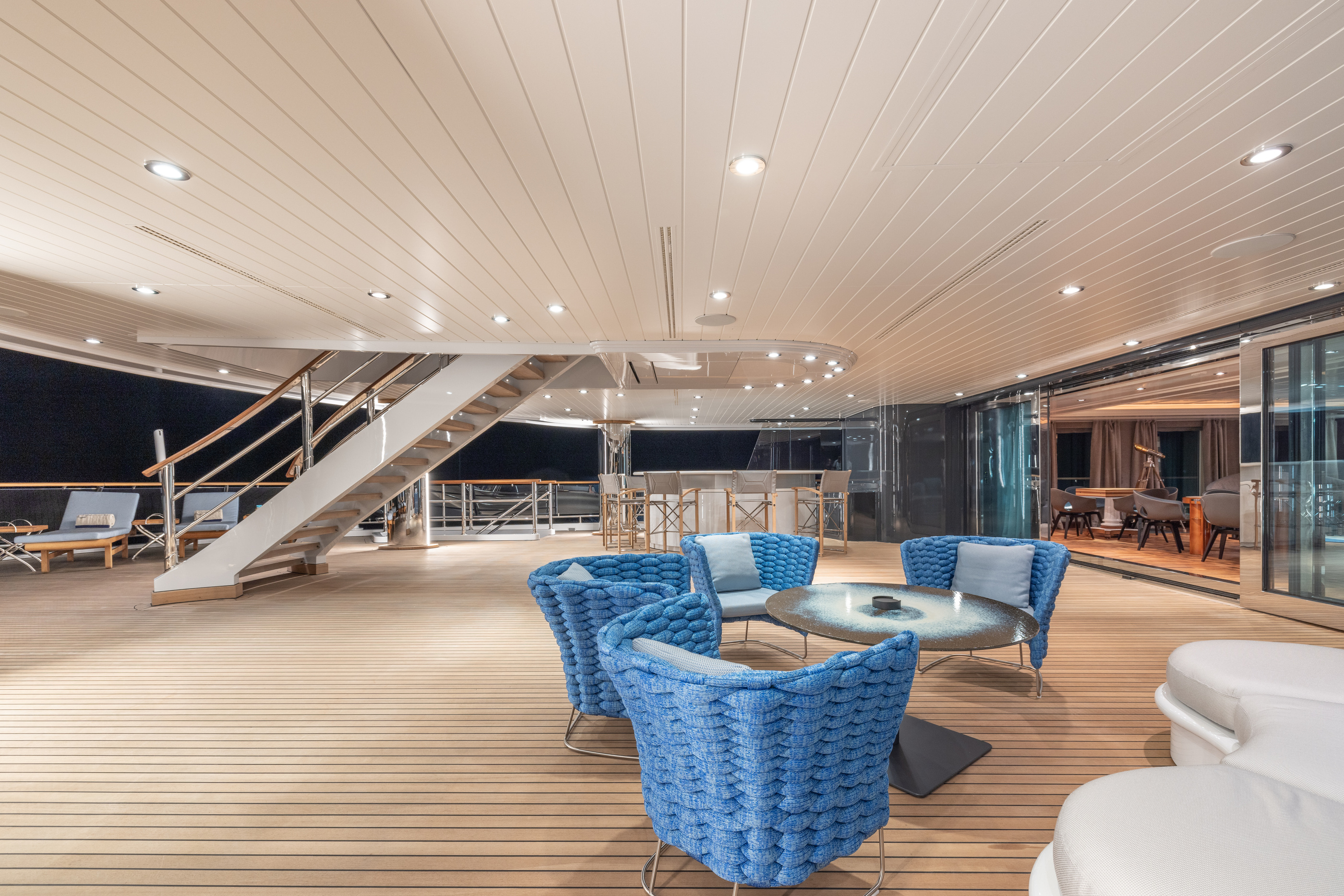 A sheltered outdoor lounge under the overhang, with the yacht’s deck stair visible behind.